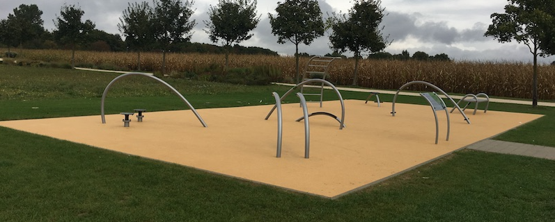 Moderner Outdoor-Fitnessbereich mit minimalistischen Geräten im Park, umgeben von Natur und Kornfeldern.