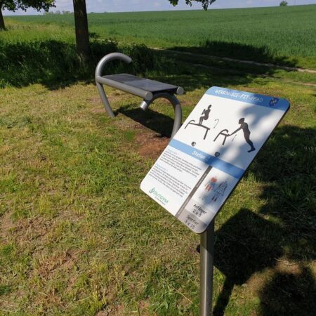 Übungsbank im Park mit Hinweisschild, umgeben von Natur und Gehweg.