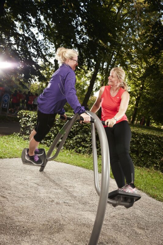 Zwei Frauen trainieren im Park an Fitnessgeräten, umgeben von grüner Natur und Sonnenlicht.