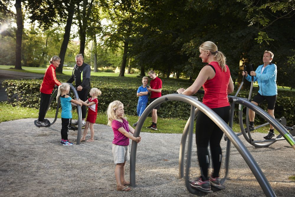 Outdoor-Fitnesspark: Menschen beim Training, Kinder aktiv, sonniger Tag, grüne Umgebung.