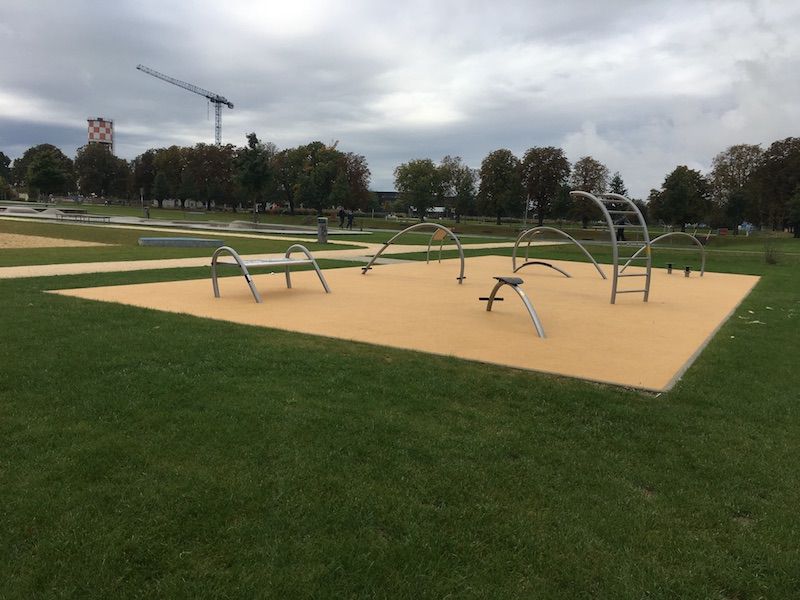 Moderner Stadtpark mit futuristischem Spielplatz, Stahlstrukturen, grüne Wiesen, Bäume, urbanes Umfeld.