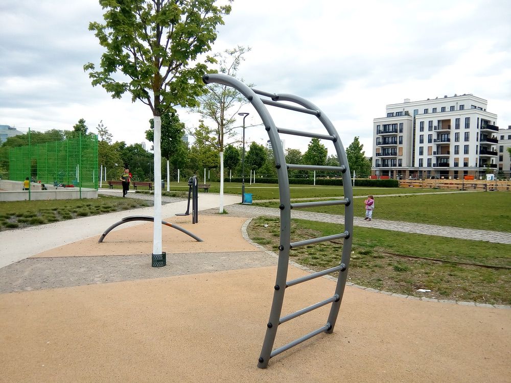 Moderner Stadtpark mit Klettergerüst, Spielplatz, grüner Umgebung und urbanem Wohngebäude im Hintergrund.