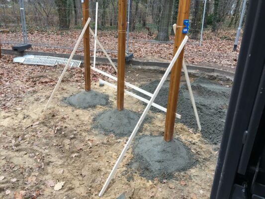 Baustelle im Waldgebiet mit frisch einbetonierten Holzpfählen für Fitnessgeräte, Herbstlaub, Sicherheitszäune.