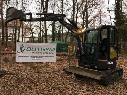 Bagger bei herbstlichem Bauprojekt im Wald für OUTGYM Fitnessumgestaltung, Bauzaun und Banner sichtbar.