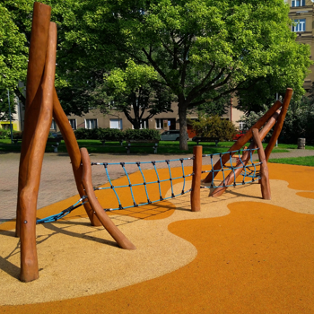 Holzspielplatz mit Netz, Gummiboden und Bäumen in urbanem Park bei sonnigem Wetter.