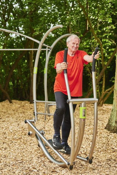 Älterer Mann trainiert im Freien auf einem elliptischen Trainer in einem grünen Park.