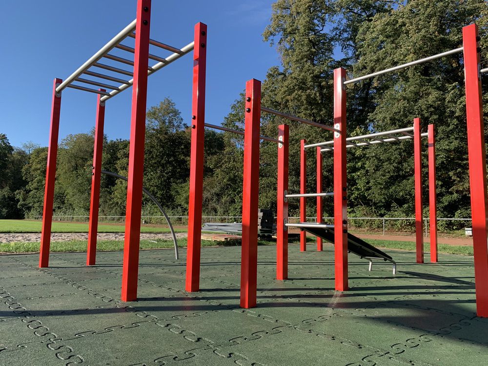 Spielplatz mit roten Klettergerüsten, Metallstangen auf grünem Sicherheitsboden, Bäume im Hintergrund.