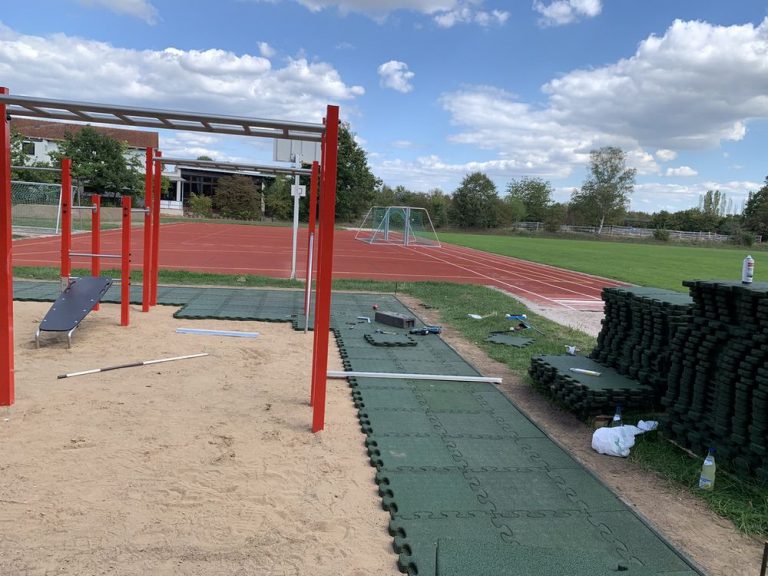 Outdoor Fitnessbereich mit Trainingsgeräten, Laufbahn und Fußballfeld im Park bei sonnigem Wetter.