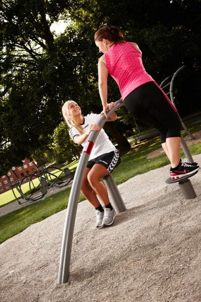 Zwei Frauen trainieren im Park auf Fitnessgeräten unter Bäumen an einem sonnigen Tag.