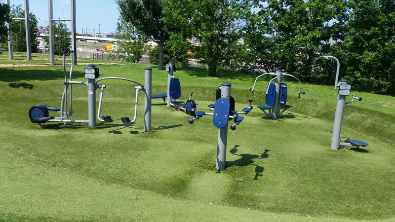 Outdoor-Fitnessbereich mit Sportgeräten auf Grünfläche, umgeben von Bäumen, urbaner Hintergrund.