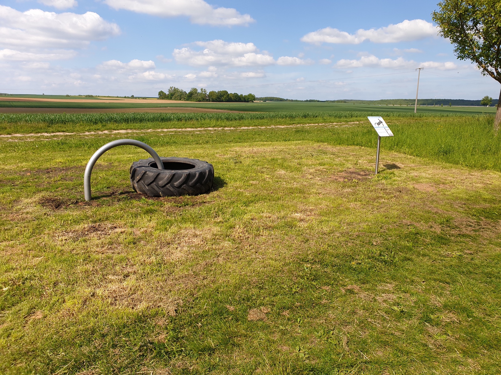 Ländliche Spielplatzszene mit Traktorreifen, Metallbögen und Feldern unter blauem Himmel.