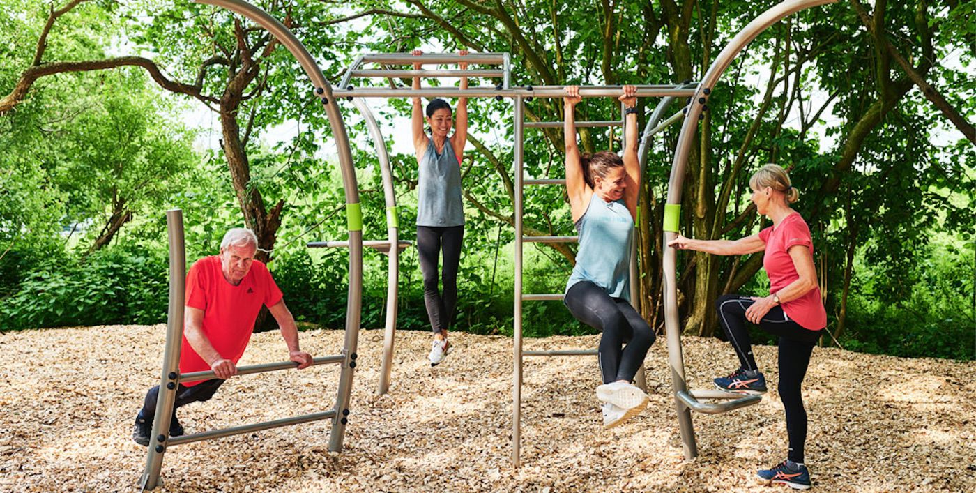 Vier Erwachsene trainieren im Freien auf Fitnessgeräten, umgeben von grüner Landschaft.