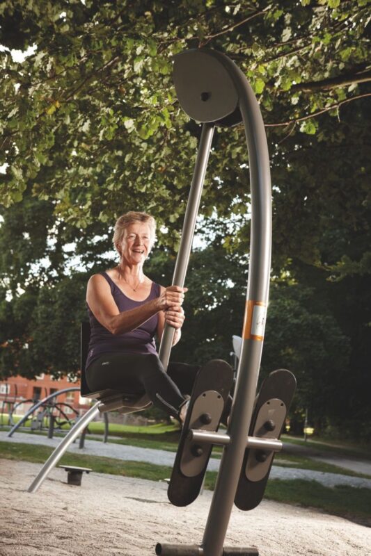 Ältere Person trainiert an moderner Outdoor-Fitnessmaschine im grünen Park.