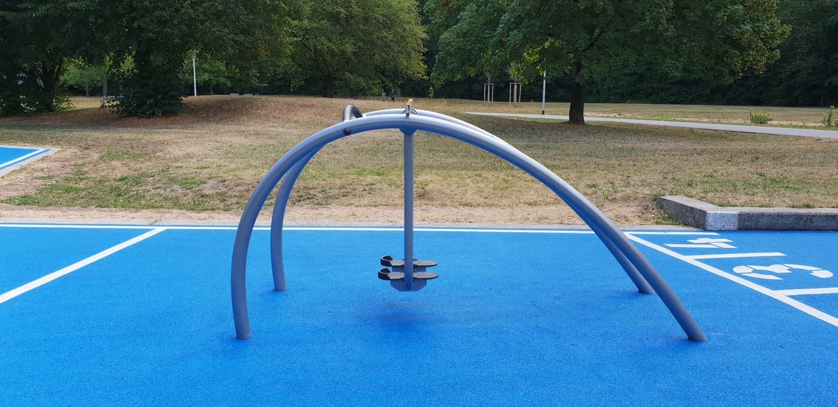 Moderner Spielplatz mit blauer Fläche, Metallkuppel, Drehsitz, umgeben von Bäumen und natürlicher Landschaft.
