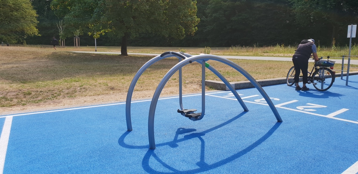 Moderner Spielplatz mit schattenwerfender Schaukel, blauem Gummiboden, Fahrrad und natürlicher Kulisse.