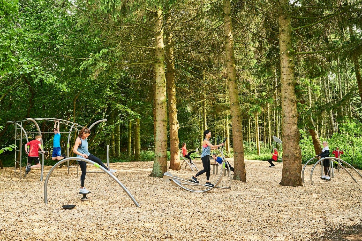 Menschen trainieren an Outdoor-Fitnessgeräten auf einer Waldfläche mit Holzhackschnitzeln.